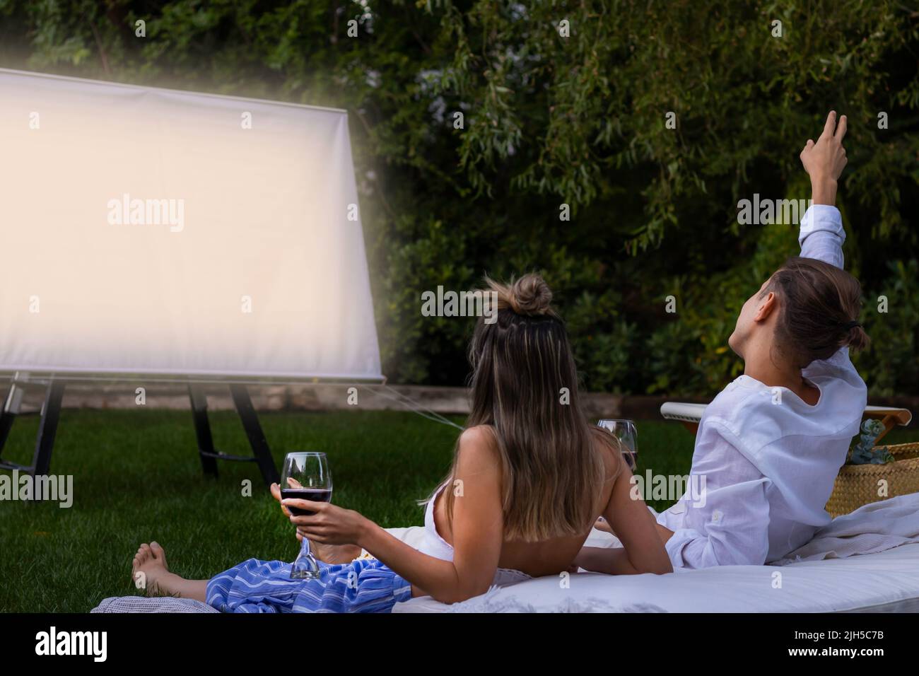 Heureux couple méconnaissable assis dans l'herbe regardant un film dans le jardin avec des verres à vin rouge Banque D'Images