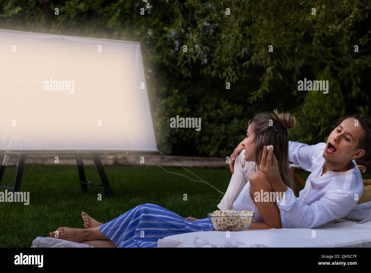 Jeune couple dans le parc regardant un film d'été à l'extérieur . Petit ami volant des opcornes de derrière à sa petite amie Banque D'Images