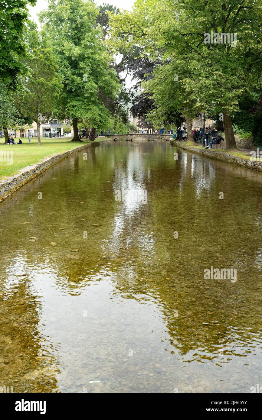 Caractéristique d'eau paisible avec des bords de pierre entourés d'arbres verdoyants et visiteurs dans le village Cotswolds Banque D'Images