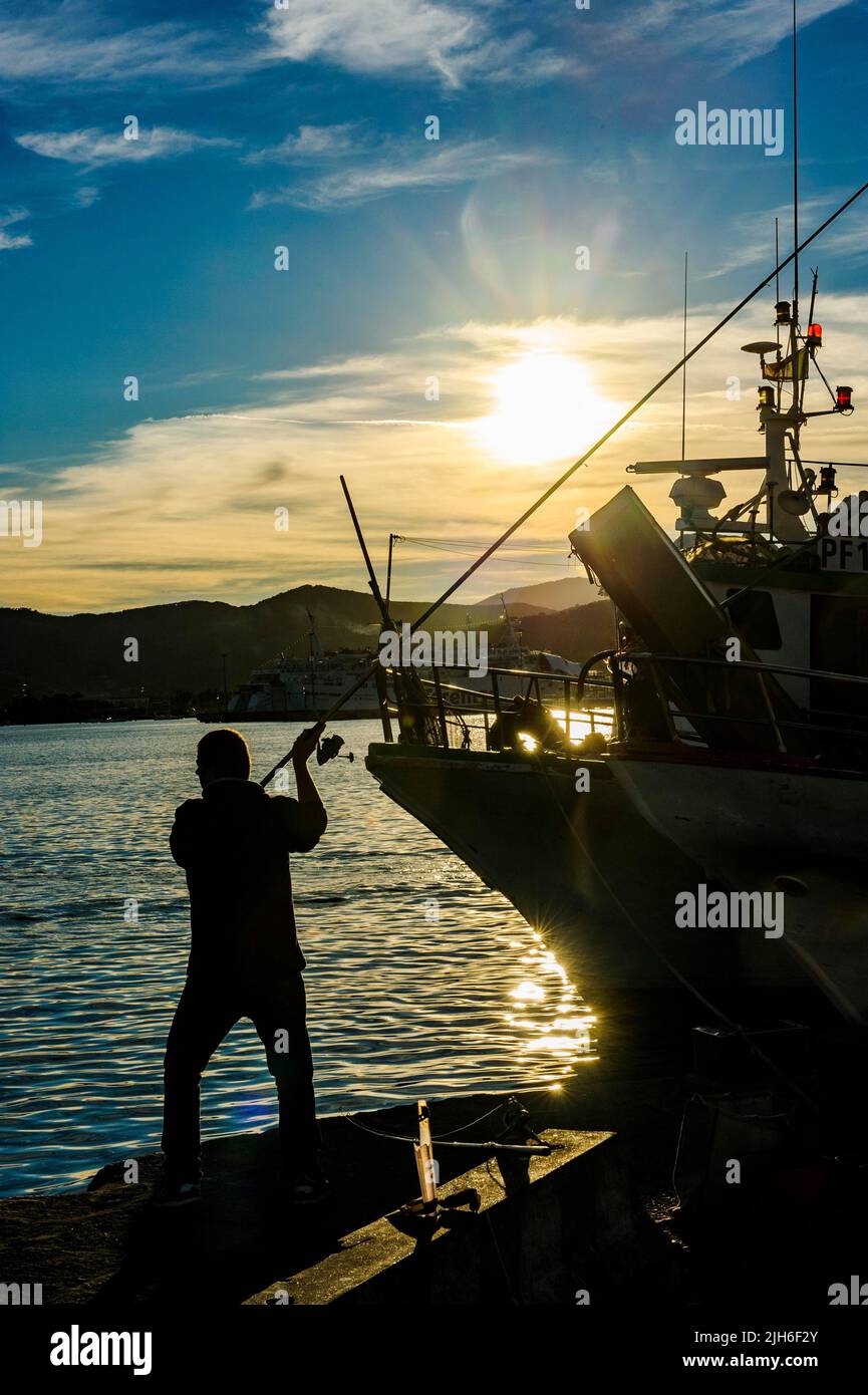 Photo de contre-jour de la silhouette de pêcheur à la ligne en casting de pêche dans le port juste en face du coucher du soleil, Portoferraio, Elba, Toscane, Italie Banque D'Images