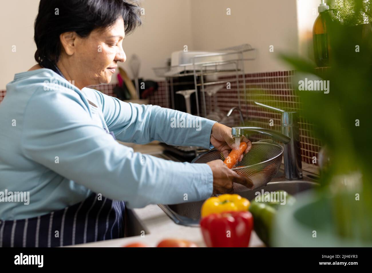 Vue latérale d'une femme biraciale mûre lavant la carotte dans la passoire sous l'eau courante dans l'évier de cuisine Banque D'Images