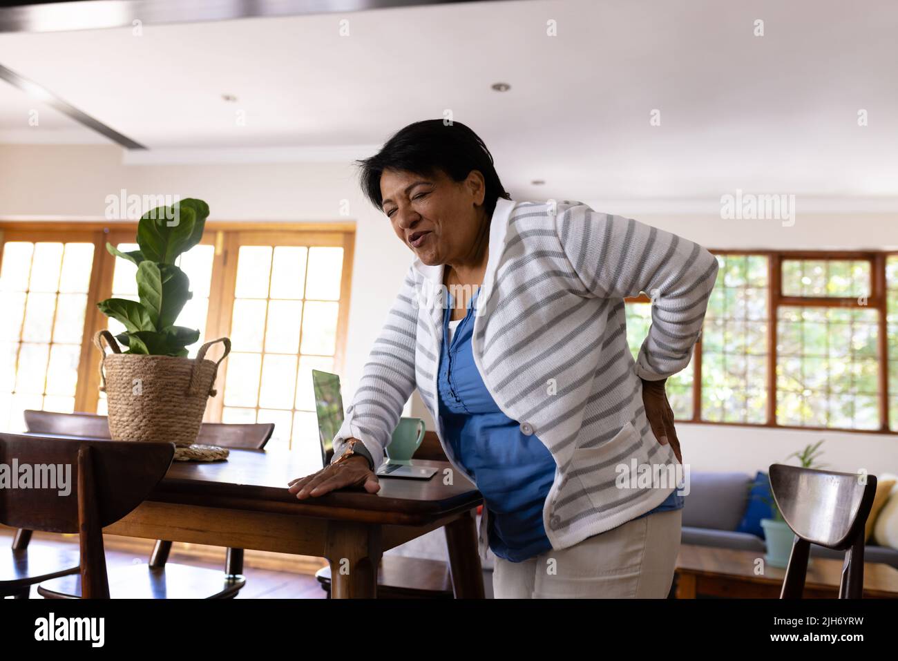 Biracial mature femme avec mal de dos touchant le dos et se tenant par une table en bois à la maison Banque D'Images