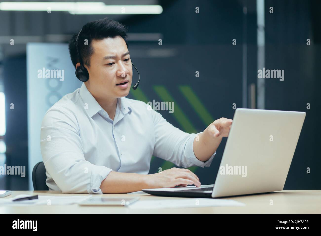 Un jeune homme d'affaires asiatique élégant dans un casque avec microphone se trouve à un ordinateur portable dans le bureau, dirige une conférence en ligne, une réunion à distance avec des clients, des partenaires. Dit, explique, sourit Banque D'Images