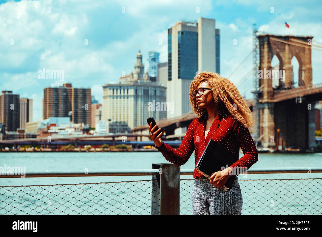 Portrait jeune femme millénaire d'entrepreneur adulte réfléchie avec des lunettes et des cheveux afro dans une conférence téléphonique vidéo à l'extérieur avec Manhattan New York City Skyline derrière le fleuve Hudson Banque D'Images