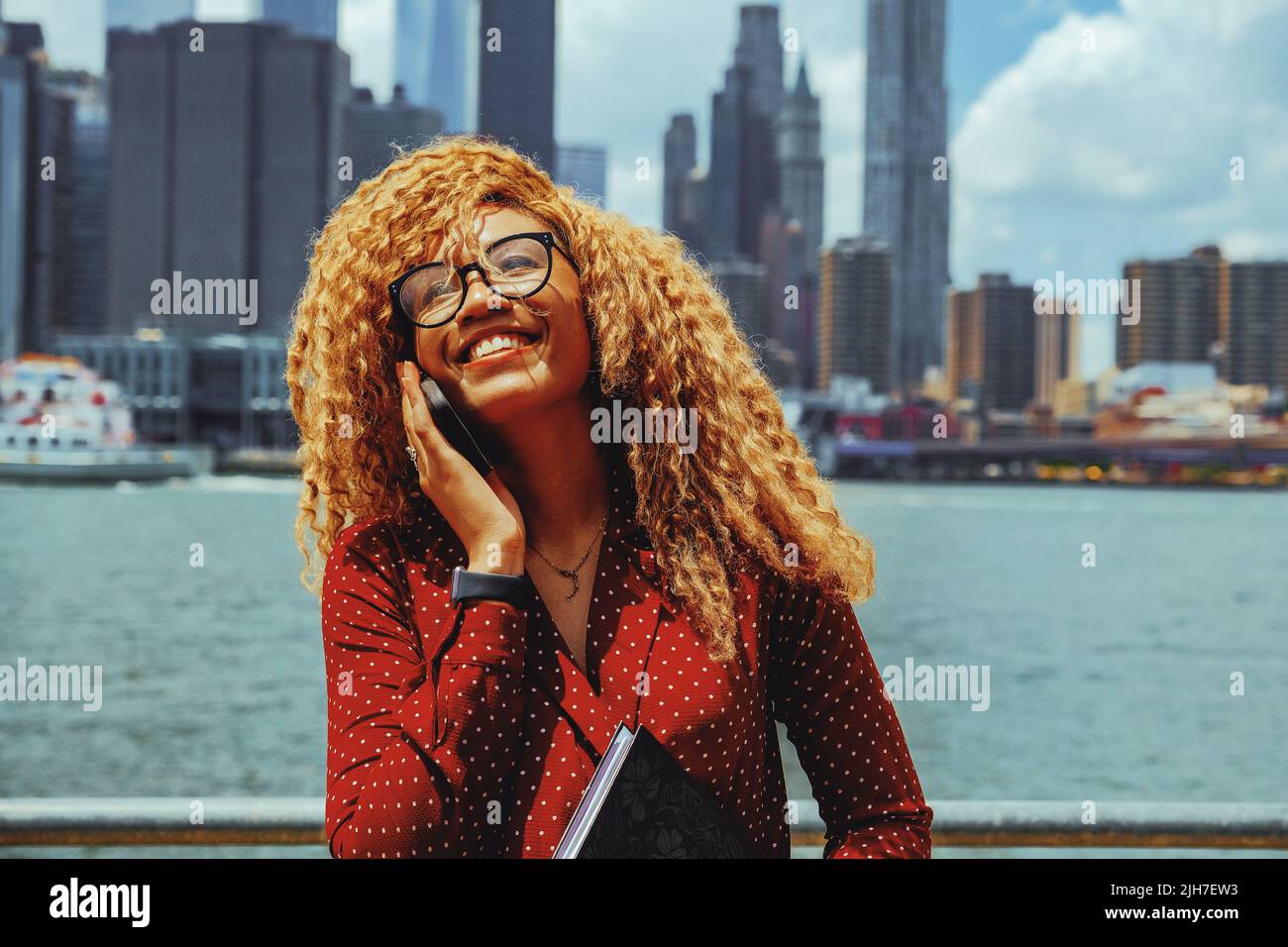 Portrait jeune entrepreneur adulte femme millénaire avec lunettes et cheveux afro souriant parlant sur un appel téléphonique à l'extérieur avec Manhattan New York City Skyline derrière Hudson River Banque D'Images