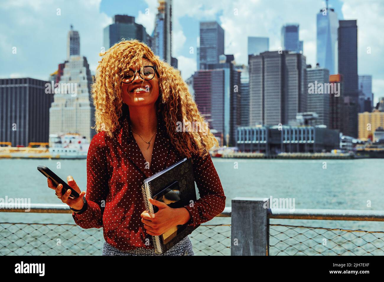 Portrait jeune entrepreneur adulte femme millénaire avec lunettes et cheveux afro souriant à l'extérieur avec Manhattan New York Skyline behindHudson River Banque D'Images