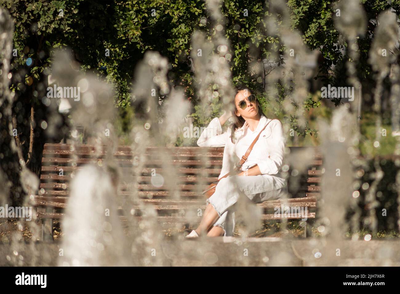 Portrait d'une jeune femme vue à travers la fontaine assis sur le banc dans le jardin de la ville, Sofia, Bulgarie, Europe de l'est, Balkans, UE Banque D'Images