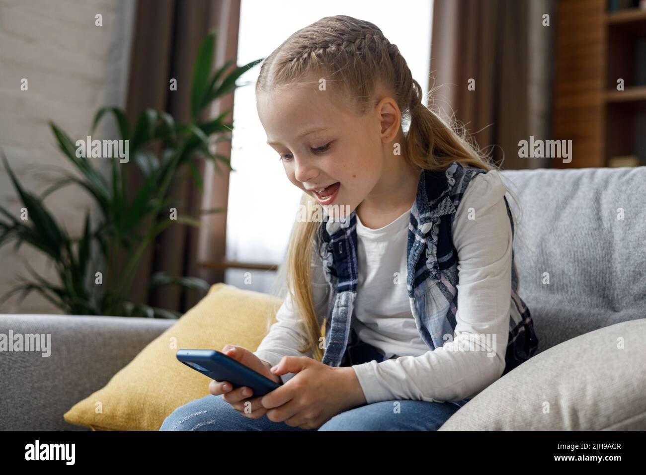 Portrait d'une petite fille mignonne tenant un smartphone et se reposant sur un canapé à la maison. Choix de musique ou de dessins animés favoris, messages texte, navigation Banque D'Images