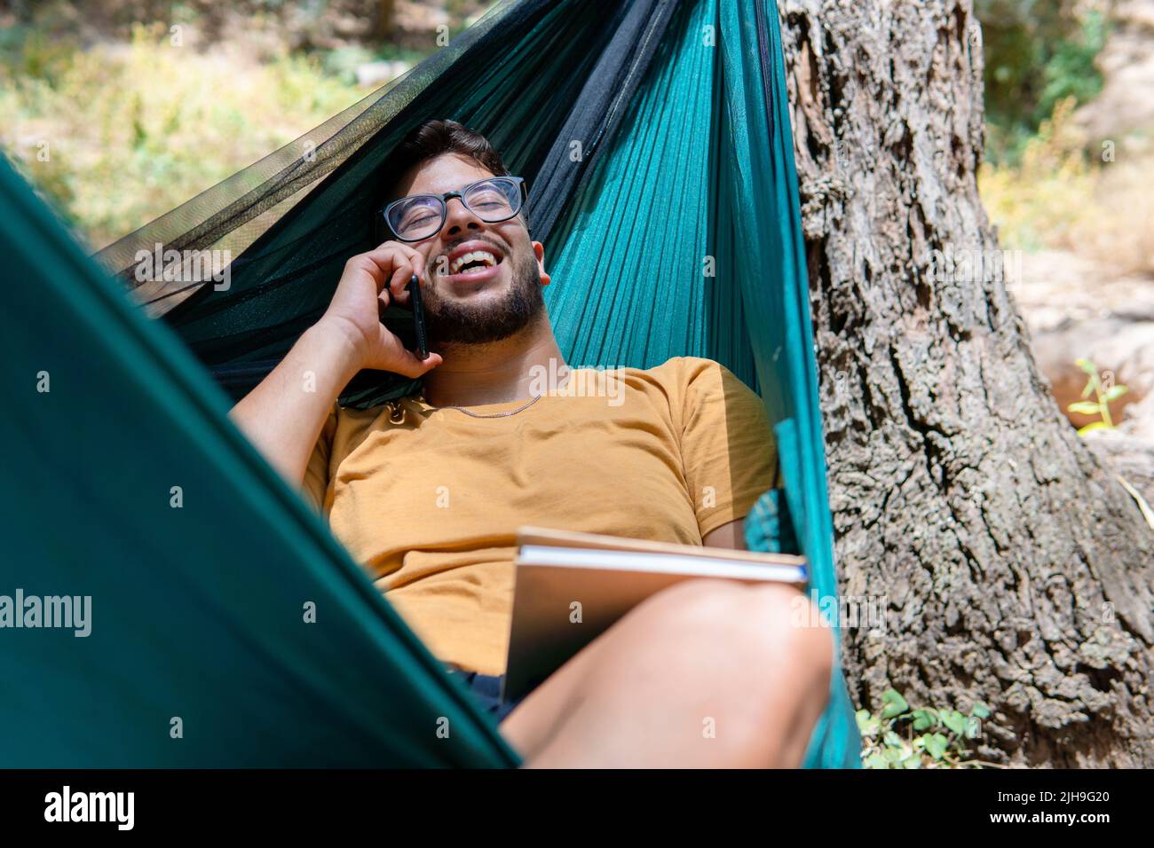 Jeune homme en vêtements et lunettes jaunes dans un hamac vert parlant au téléphone appréciant la conversation tenant un livre dans la nature Banque D'Images