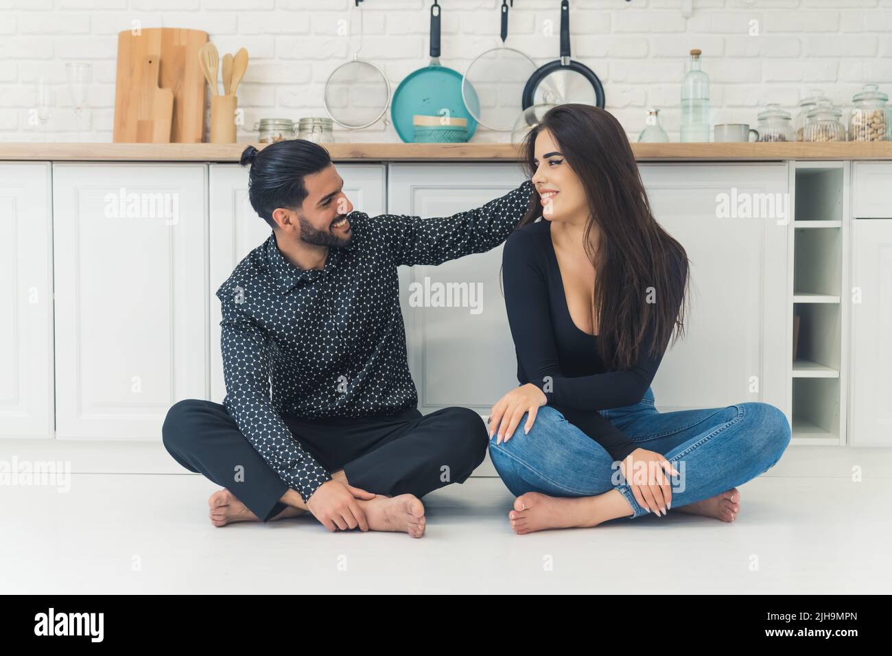 Proximité et sensibilité. Couple hétérosexuel pieds nus - barbu officiellement habillé beau homme cubain et femme caucasienne à cheveux longs - assis sur le plancher blanc de la cuisine. Homme touchant les cheveux de sa jeune femme. Cuisine intérieure moderne. Photo de haute qualité Banque D'Images