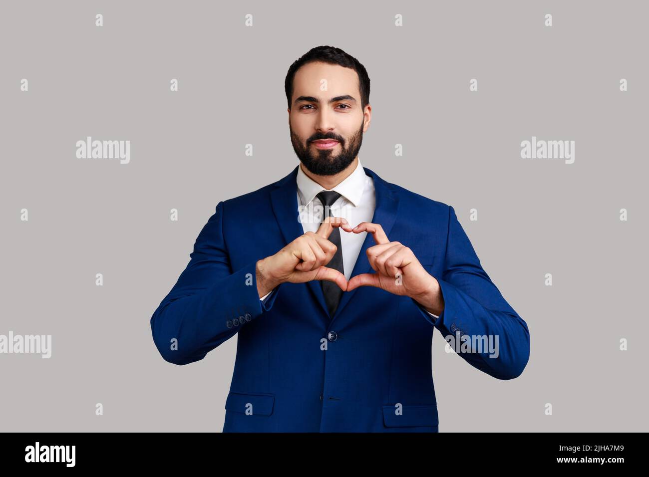 Portrait d'homme barbu romantique debout avec le geste de main d'amour sur le coeur et regardant l'appareil photo souriant, portant le costume de style officiel. Prise de vue en studio isolée sur fond gris. Banque D'Images