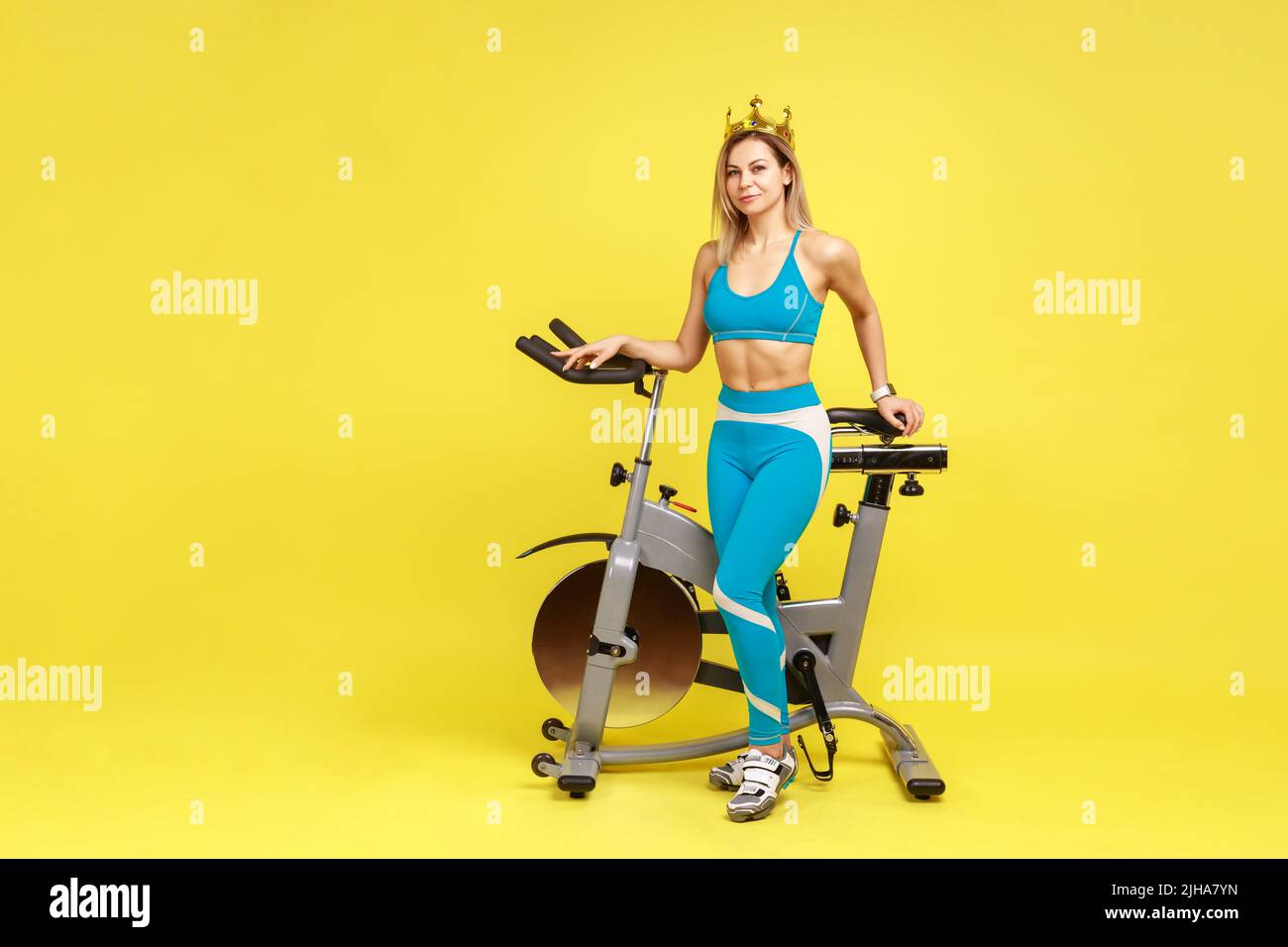 Portrait en longueur d'une femme pleine d'assurance près d'un équipement de sport avec couronne dorée sur sa tête, portant des vêtements de sport bleus. Studio d'intérieur isolé sur fond jaune. Banque D'Images