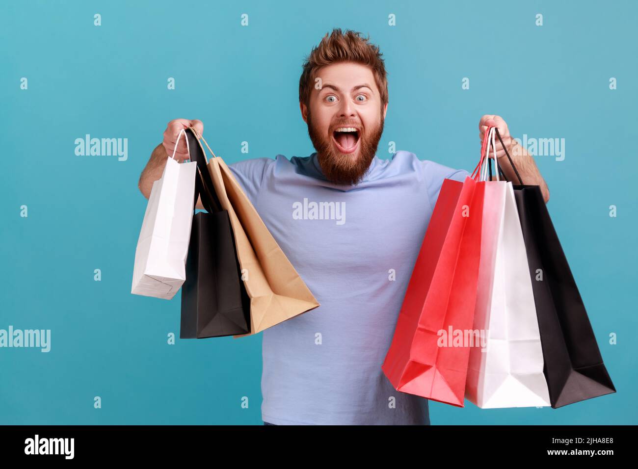 Portrait d'un bel homme barbu excité et stupéfié tenant des sacs de papier de shopping, heureux et heureux avec des réductions de centre commercial, de bons achats. Studio d'intérieur isolé sur fond bleu. Banque D'Images