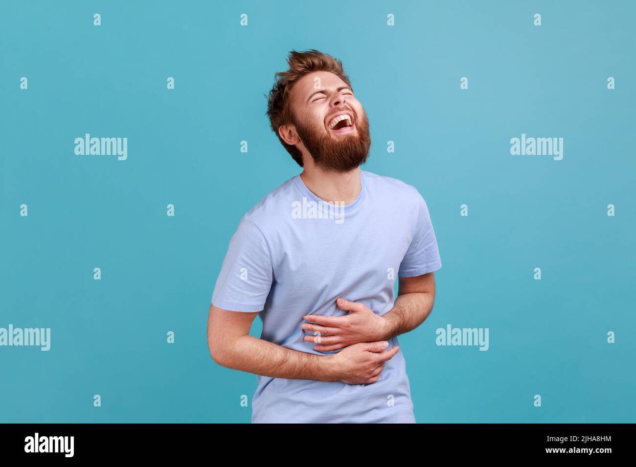 Portrait d'un homme barbu joyeux et positif en riant avec joie à quelque chose garde les mains sur le ventre, sourit globalement, exprimant des émotions positives. Studio d'intérieur isolé sur fond bleu. Banque D'Images