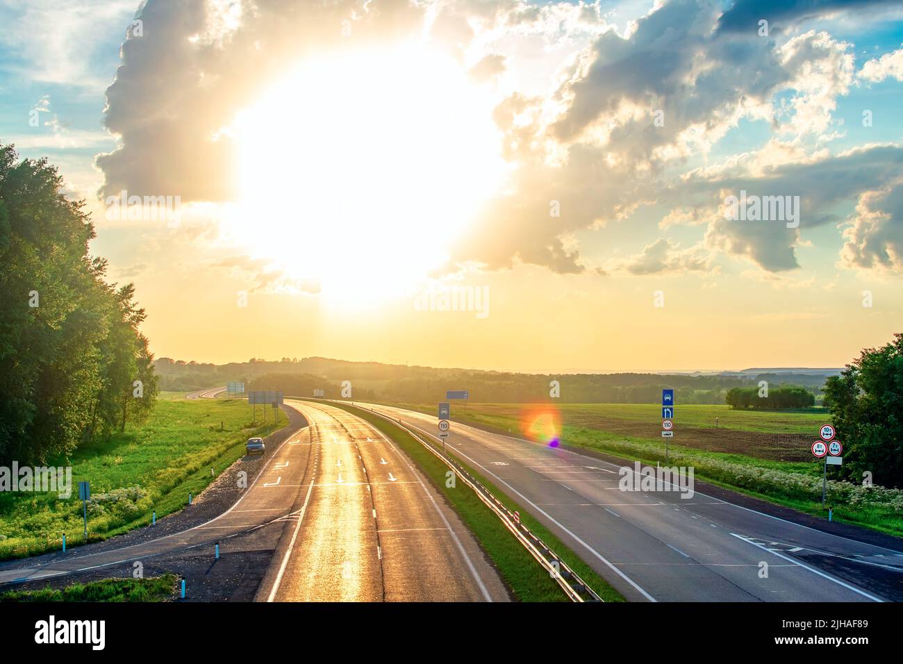 paysage d'été, autoroute avec des panneaux de signalisation qui brillent avec la lumière réfléchie avec l'éblouissement du soleil orange lumineux et aveuglant de coucher de soleil Banque D'Images