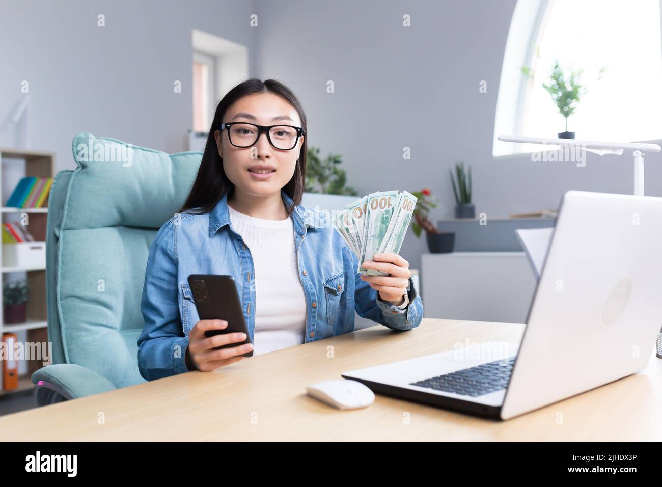 Formation professionnelle sur l'argent. Une jeune femme d'affaires asiatique organise une session de formation sur le thème de l'argent. Il tient un téléphone et une liade de factures. Assis à son bureau, parle, regarde dans la caméra. Banque D'Images