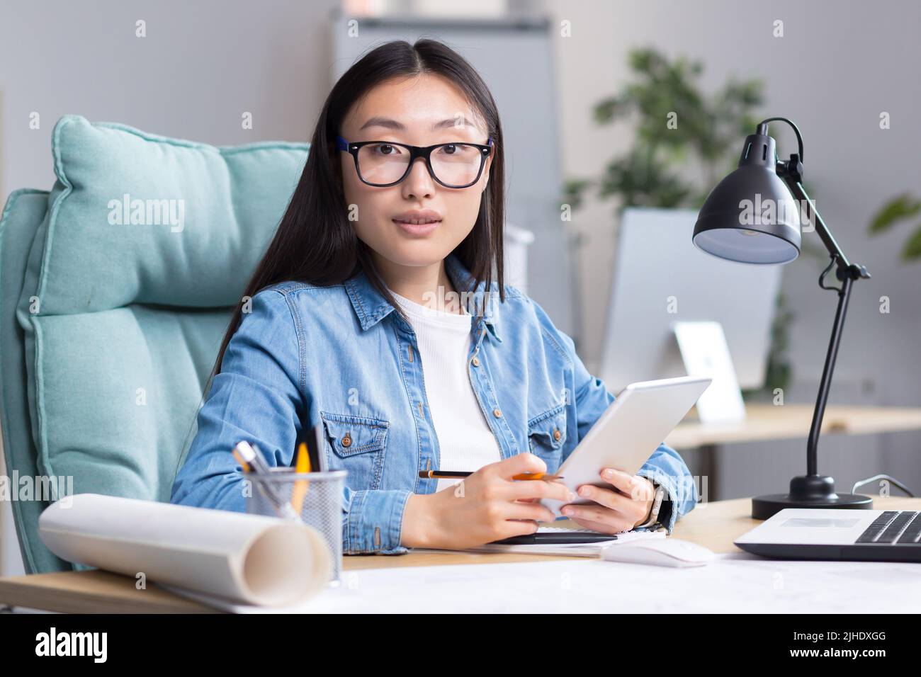 Portrait d'une jeune belle architecte asiatique-designer femme en lunettes travaille sur un projet, assis à un bureau, tient une tablette dans ses mains. Il regarde la caméra, sourit. Banque D'Images