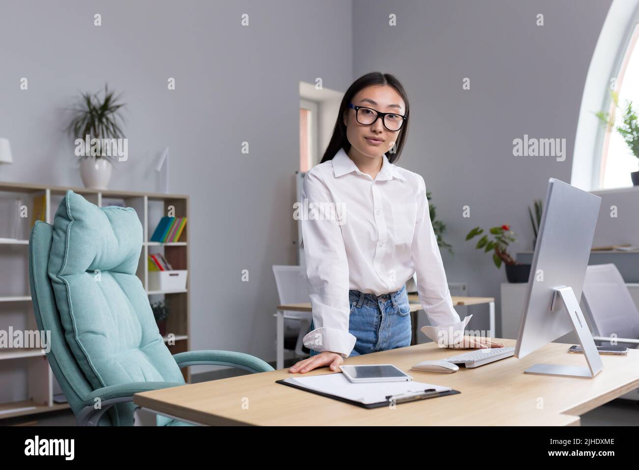 Portrait d'une jeune femme asiatique en lunettes femme d'affaires, gestionnaire, directeur, patron debout dans le bureau au bureau. Elle se penche sur la table avec ses mains, regarde la caméra, sourit. Banque D'Images