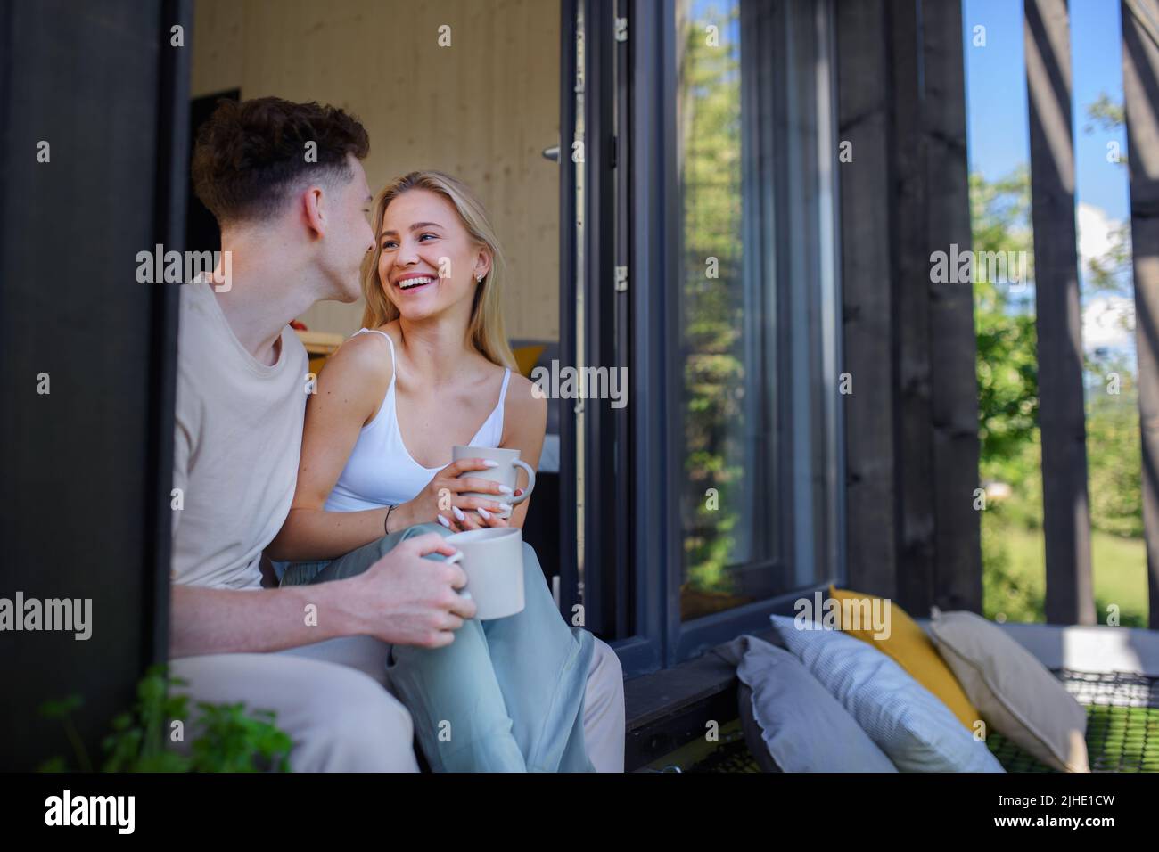 Jeune couple assis, buvant du café et des câlins dans une terrasse hamac dans leur nouvelle maison dans une petite maison en bois, concept de vie durable. Banque D'Images