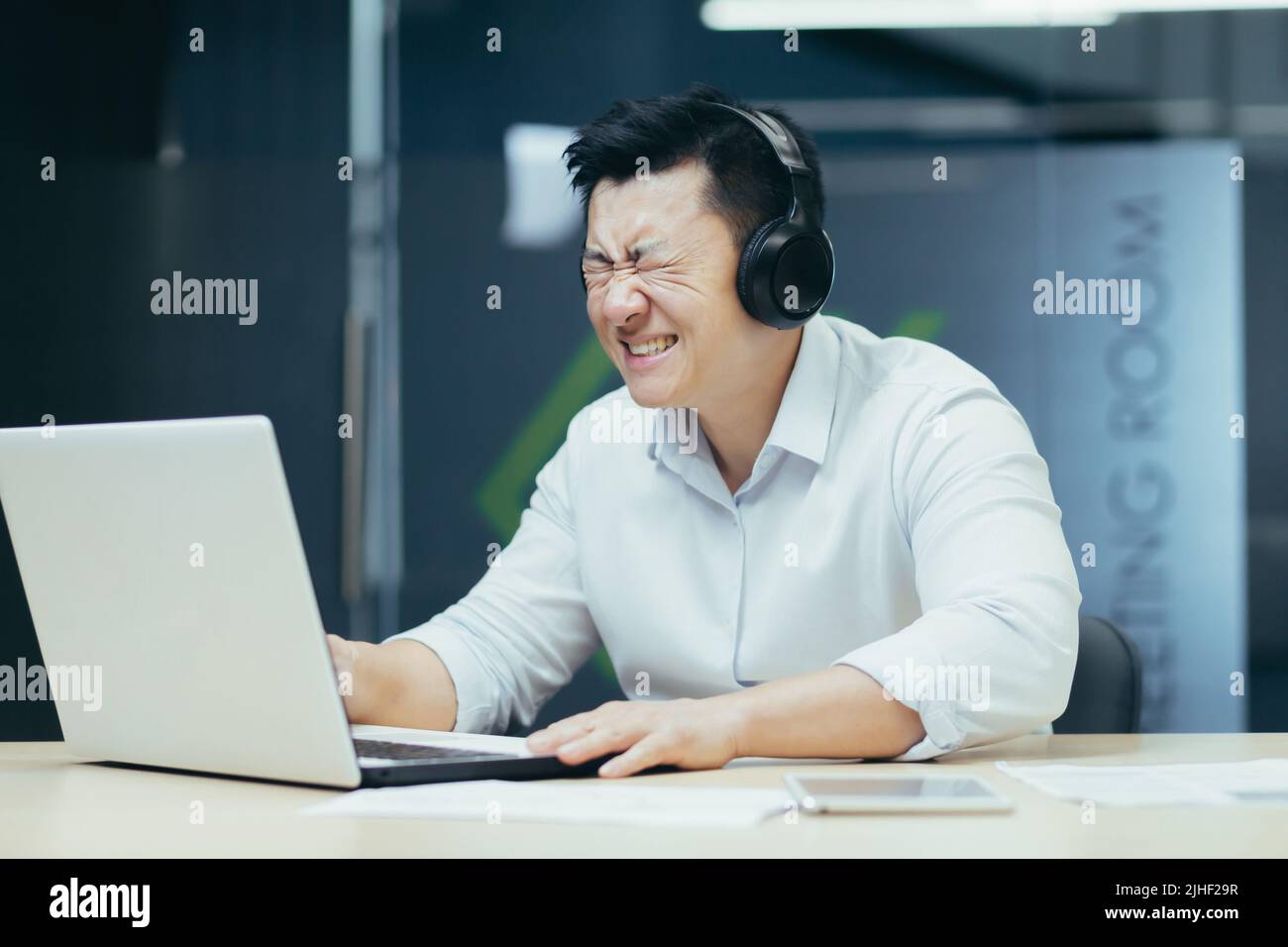 Homme d'affaires asiatique regardant un match de sport au bureau, homme regardant déçu par l'ordinateur portable, utilisant des écouteurs pour écouter Banque D'Images