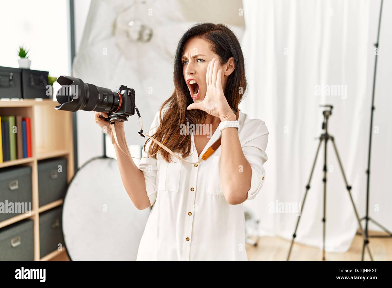 Belle femme caucasienne travaillant comme photographe au studio de photographie criant et criant à voix haute à côté de la main sur la bouche. Concept de communication. Banque D'Images