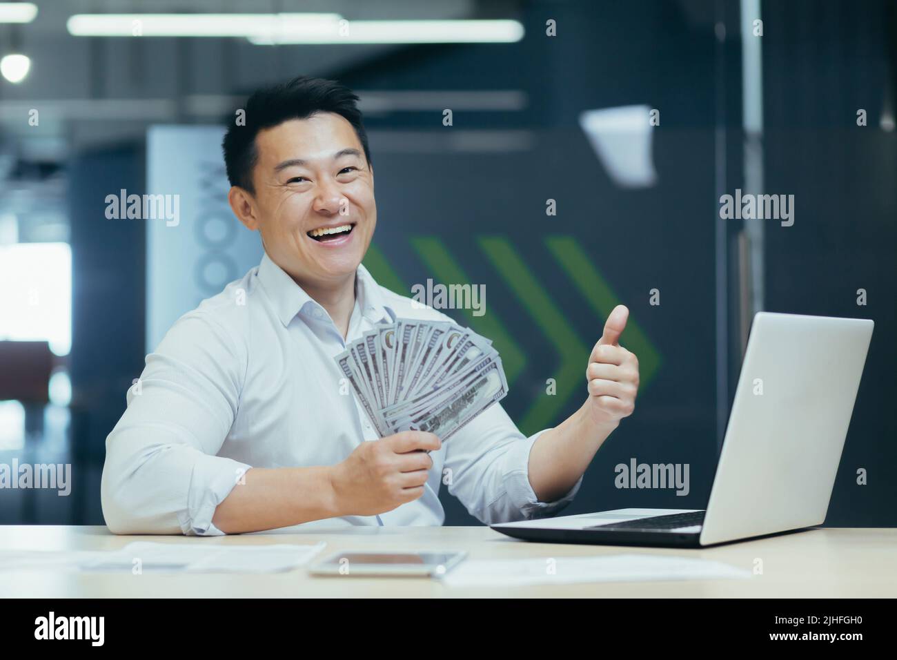 Portrait d'un homme d'affaires asiatique heureux au bureau avec des dollars d'argent liquide, homme souriant et regardant l'appareil photo, tenant des dollars à la main et pouces vers le haut Banque D'Images