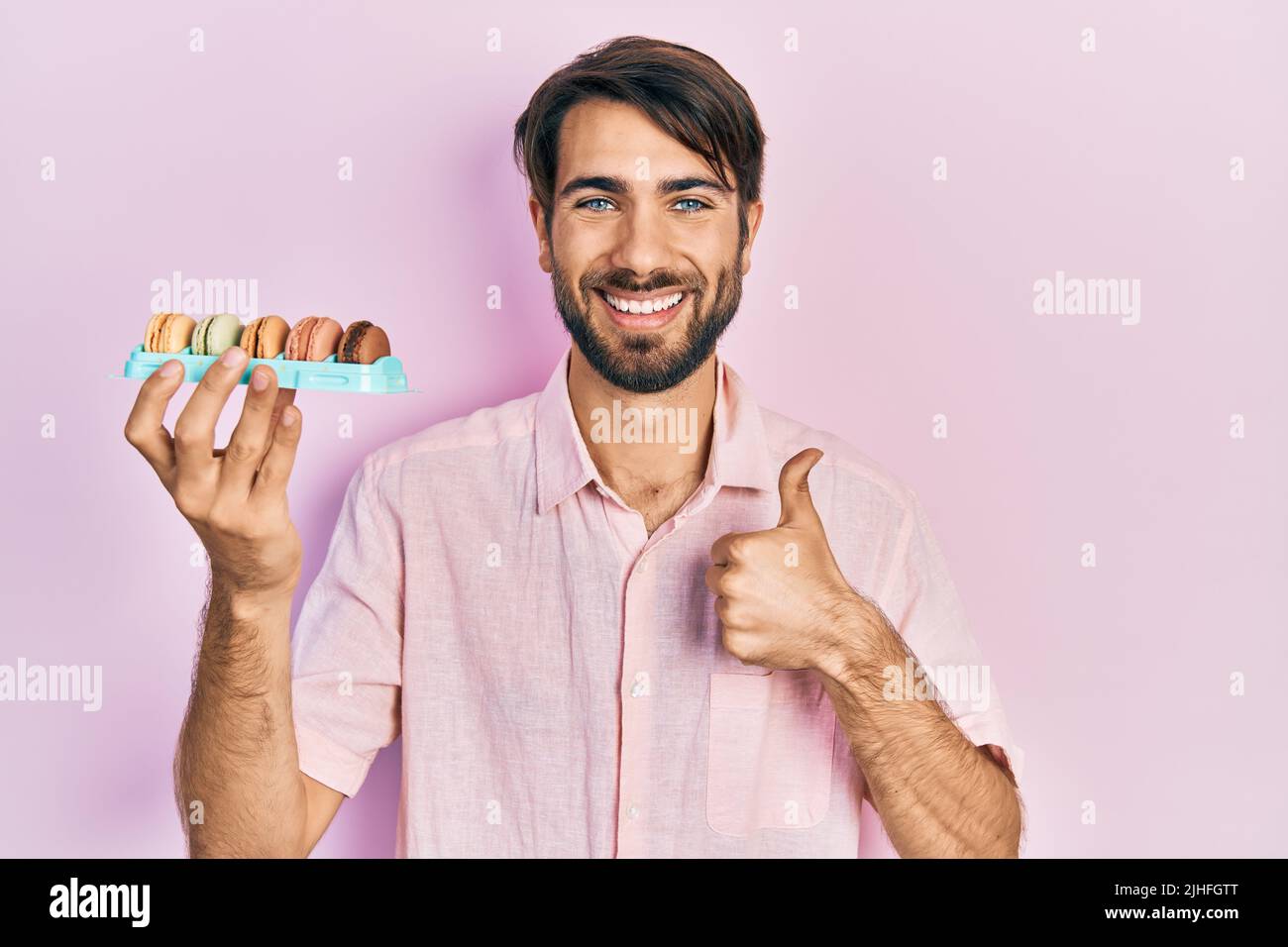 Jeune homme hispanique tenant de délicieux macrons pâtisseries souriant heureux et positif, pouce vers le haut faisant excellent et signe d'approbation Banque D'Images