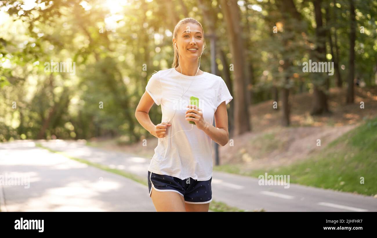 Femme écouter de la musique courir sur la piste d'asphalte parc d'été Caucasien Ecouteurs femelles jogging matin ensoleillé entraînement de coureur tenir le smartphone dans Talon de la main Banque D'Images