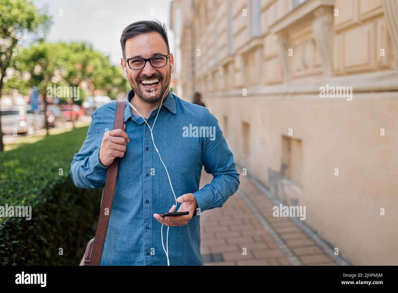 Un jeune homme d'affaires heureux utilisant un casque. Un homme de banlieue professionnel écoute de la musique depuis un smartphone. Il est debout sur le trottoir par des plantes dans le Banque D'Images
