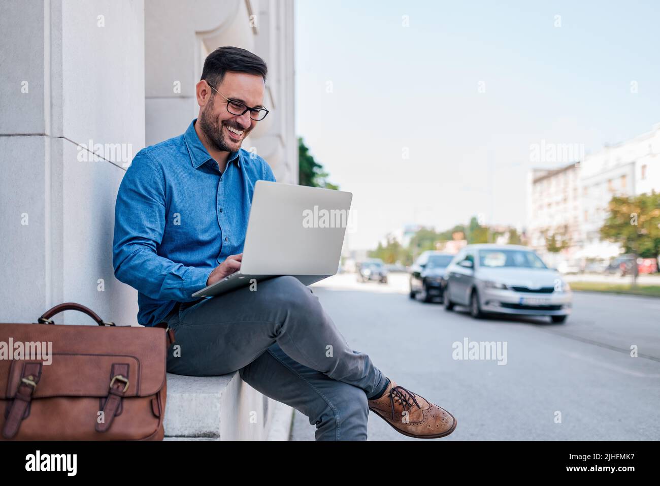 Portrait d'un entrepreneur heureux tenant et travaillant sur un ordinateur portable tout en étant assis dans la rue urbaine. Jeune homme d'affaires élégant portant un sac pour ordinateur portable et moi Banque D'Images