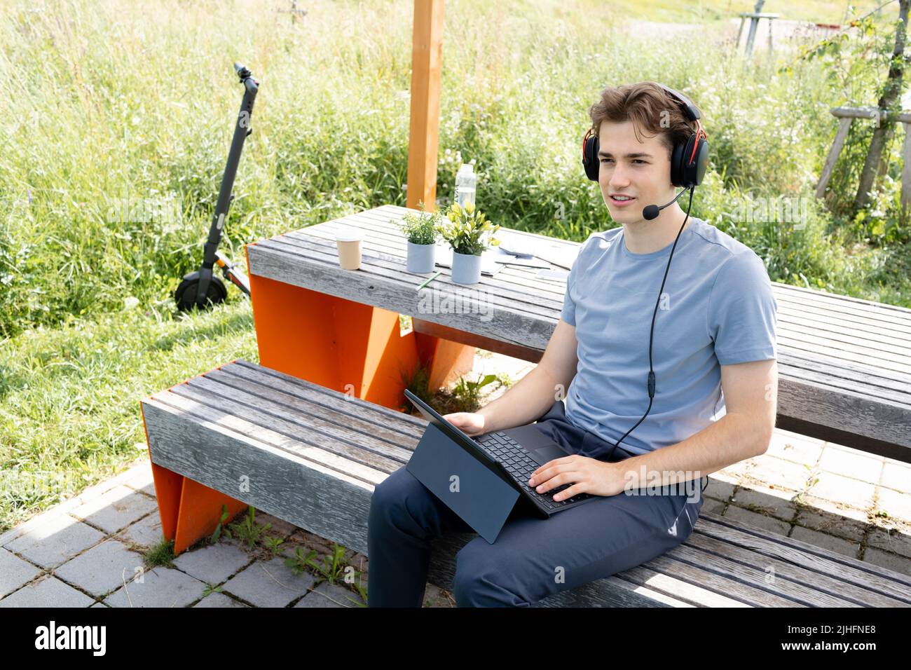 cool, jeune homme avec chemise grise assis à l'extérieur dans un parc avec casque et de travail sur sa tablette, en arrière-plan e-scooter est debout Banque D'Images