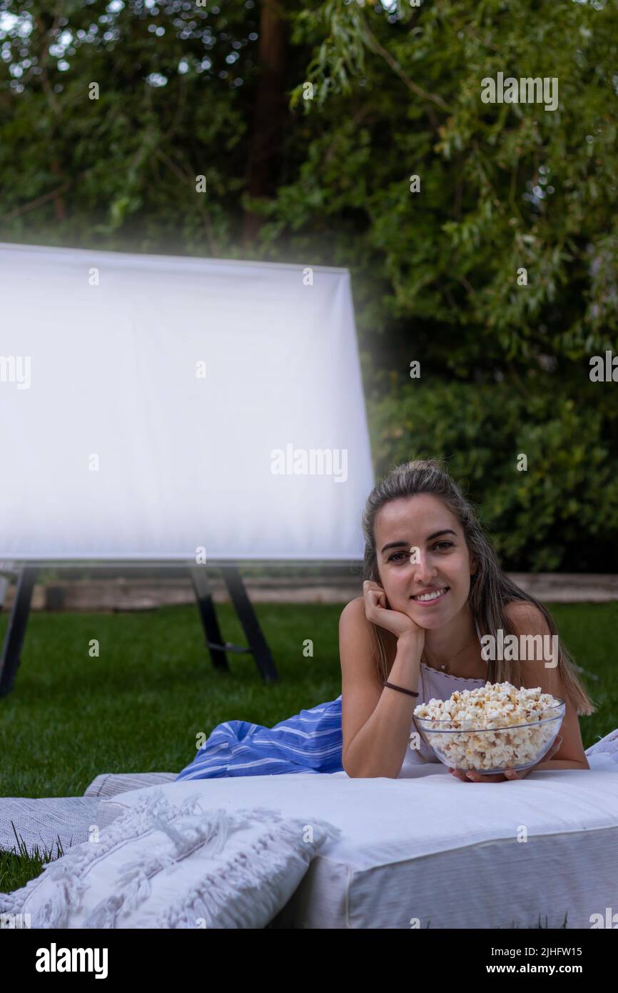 Vue verticale de la jeune femme regardant la caméra sourire avec un écran de projecteur en arrière-plan et quelques popcorns Banque D'Images