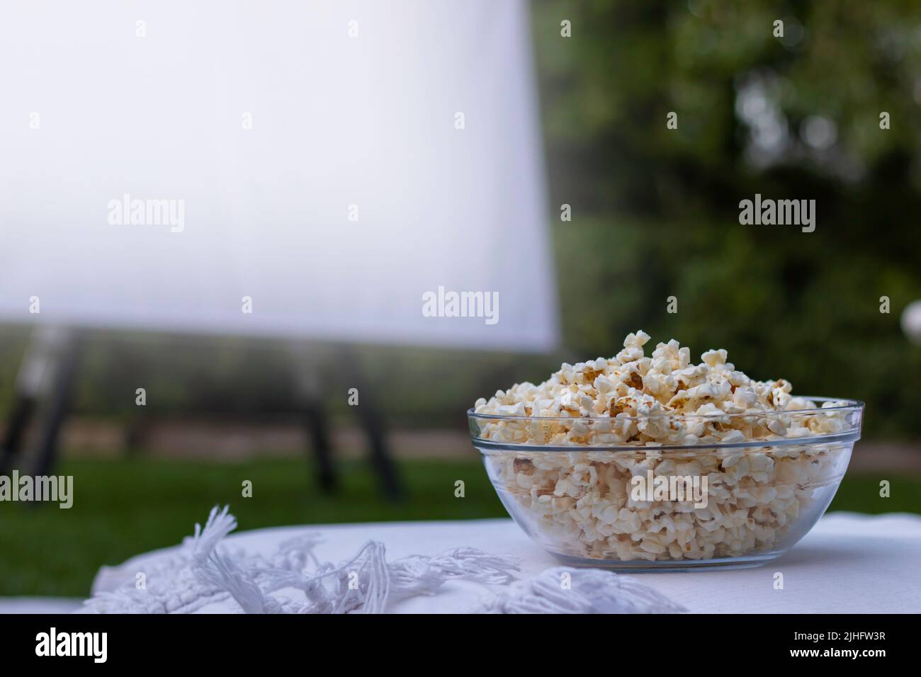 Gros plan sur un bol de pop-corn transparent avec un écran de projecteur dans un arrière-plan flou au cinéma d'été en plein air Banque D'Images