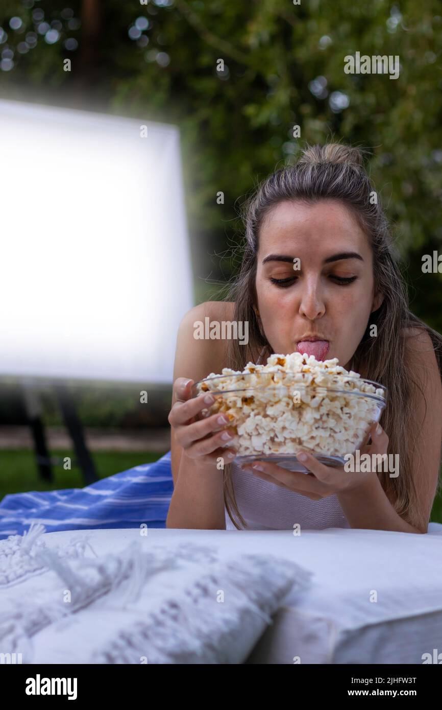 Jeune belle femme avec la bouche dehors léchant un bol de popcorns pendant que dans le cinéma d'été en plein air dans le parc Banque D'Images