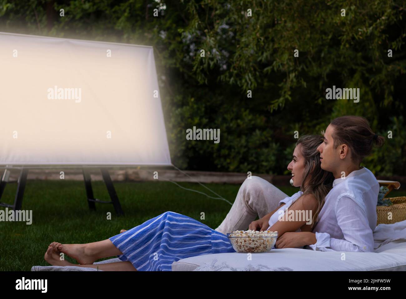 Un jeune couple mignon qui s'assoit dans le jardin pendant une nuit de nuit en regardant un film dans le cinéma en plein air en été Banque D'Images