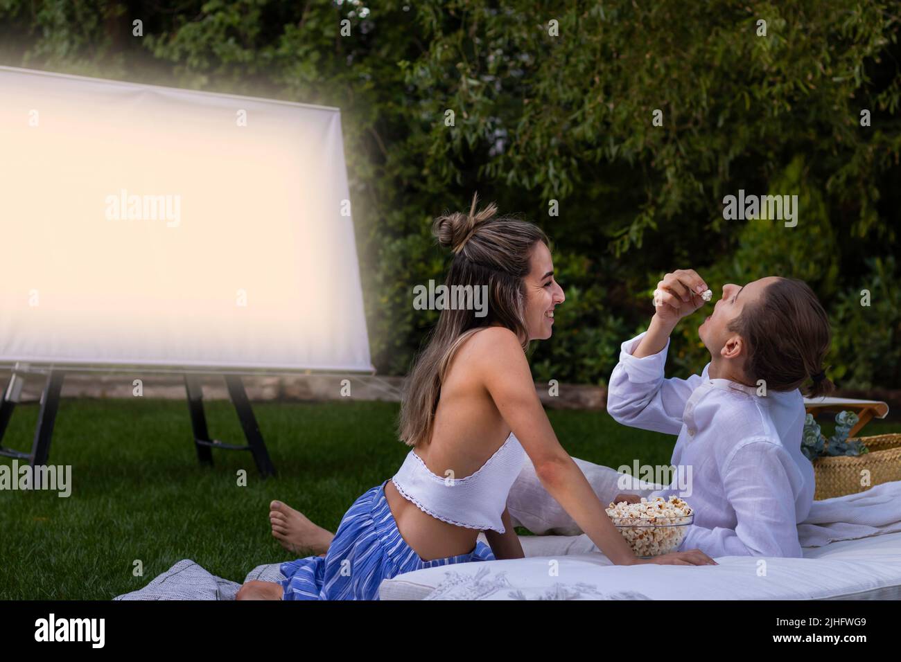Un jeune couple mignon qui interagit souriant et mangeant des popcorns tout en regardant un film dans le jardin la nuit Banque D'Images