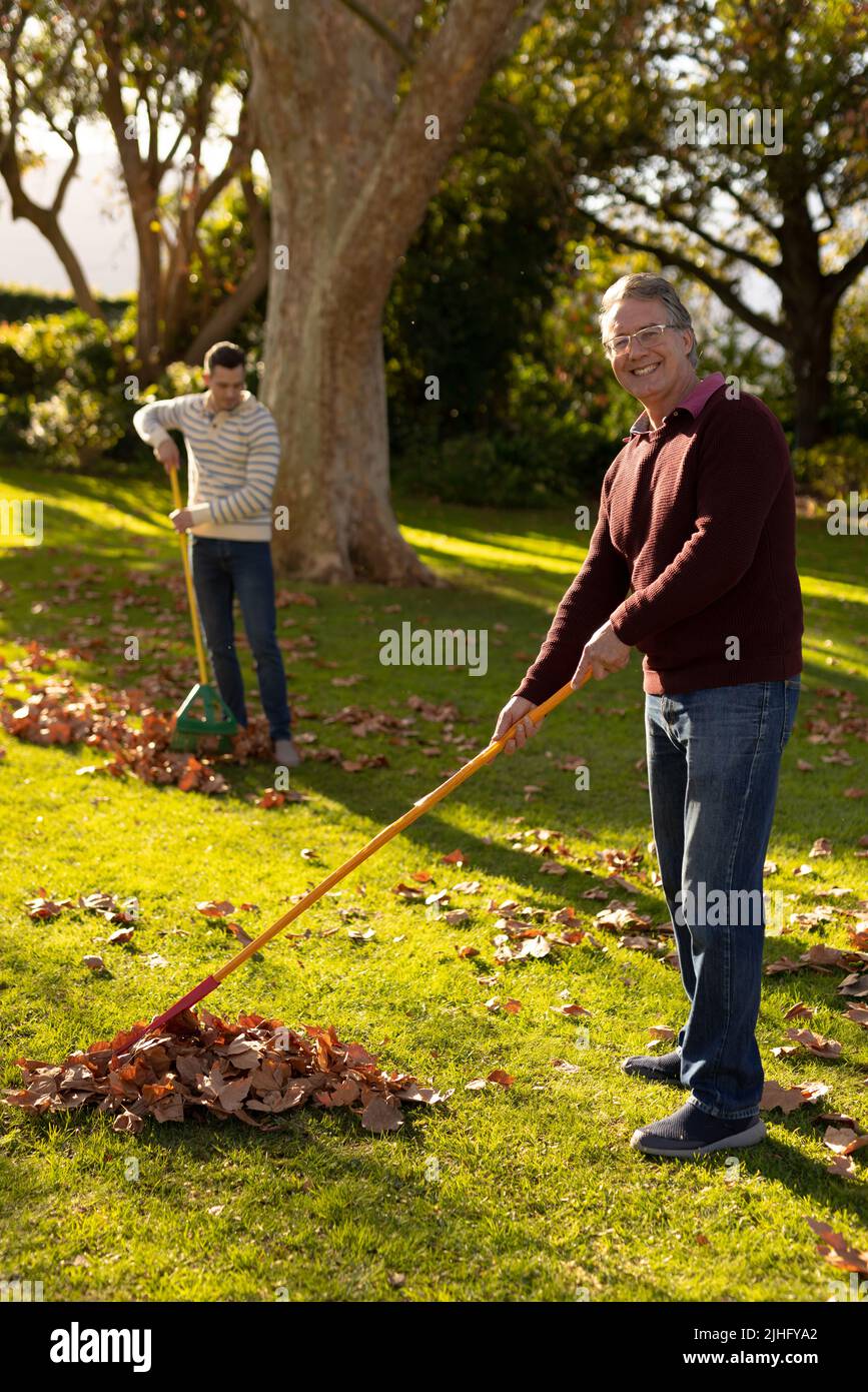 Image verticale de père caucasien heureux et de fils adultes qui balaie les feuilles dans le jardin Banque D'Images