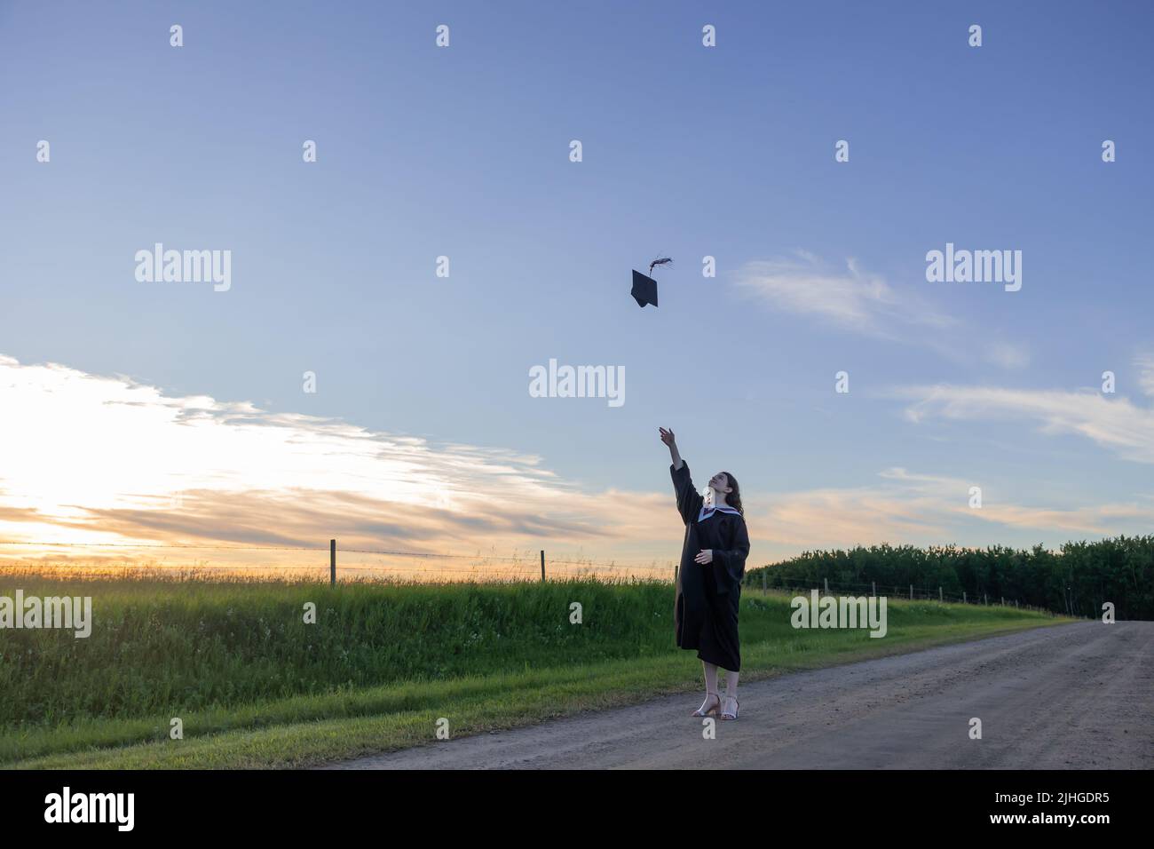 Une jeune femme portant une robe de graduation jette sa casquette, un plateau de mortier, dans l'air avec un ciel bleu à l'arrière-plan Banque D'Images