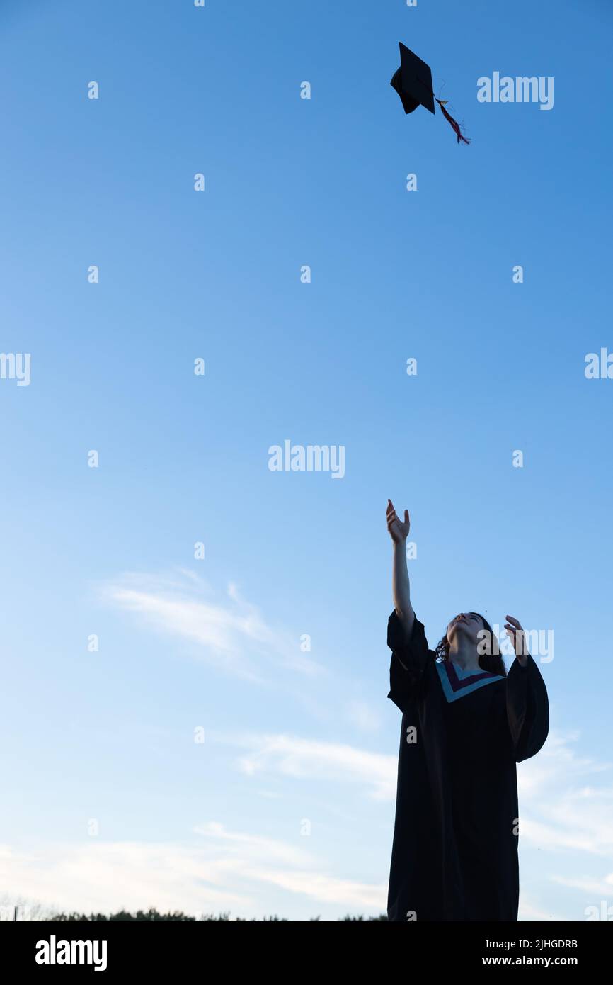 Une jeune femme portant une robe de graduation jette sa casquette, un plateau de mortier, dans l'air avec un ciel bleu à l'arrière-plan Banque D'Images