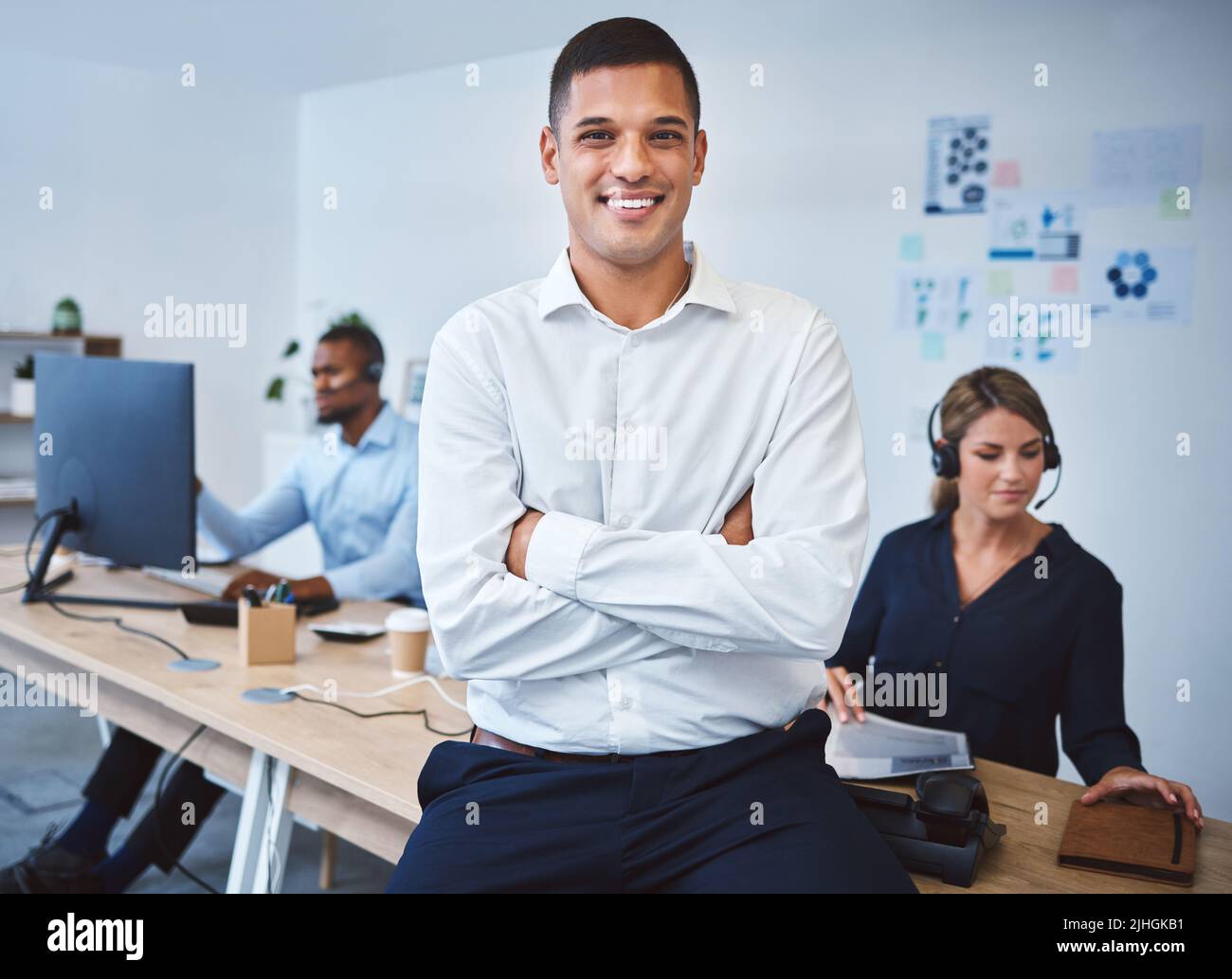 Portrait d'un jeune agent de télémarketing de centre d'appels de course mixte confiant debout avec les armes croisées tout en travaillant dans un centre d'appels avec des collègues dans Banque D'Images