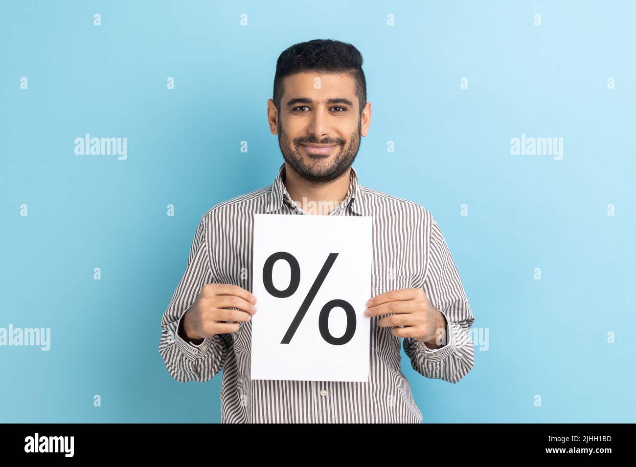 Homme d'affaires satisfait positif tenant le papier et montrant avec l'inscription de signe de pourcentage, regardant la caméra avec le sourire, portant une chemise rayée. Studio d'intérieur isolé sur fond bleu. Banque D'Images