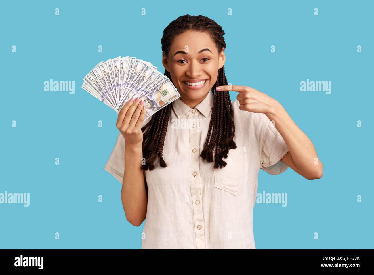 Femme souriante positive avec des dreadlocks noirs tenant des billets de banque en dollars, pointant de l'argent, regardant l'appareil photo avec une expression heureuse, portant une chemise blanche. Studio d'intérieur isolé sur fond bleu. Banque D'Images
