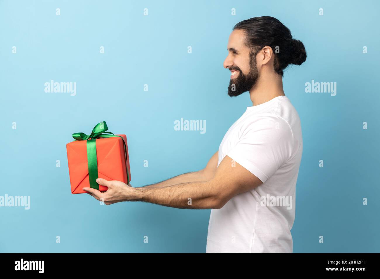 Vue latérale d'un bel homme enchanté avec une barbe portant un T-shirt blanc donnant une boîte cadeau enveloppée et souriant, regardant à l'avance, des vacances heureuses. Studio d'intérieur isolé sur fond bleu. Banque D'Images