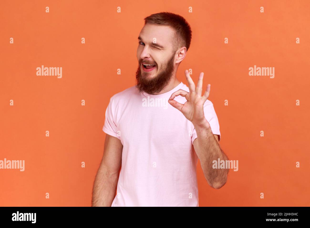 Portrait de l'homme barbu debout avec les pouces vers le haut, comme un geste, démontrant l'approbation et d'accord avec la suggestion, portant le T-shirt rose. Studio d'intérieur isolé sur fond orange. Banque D'Images