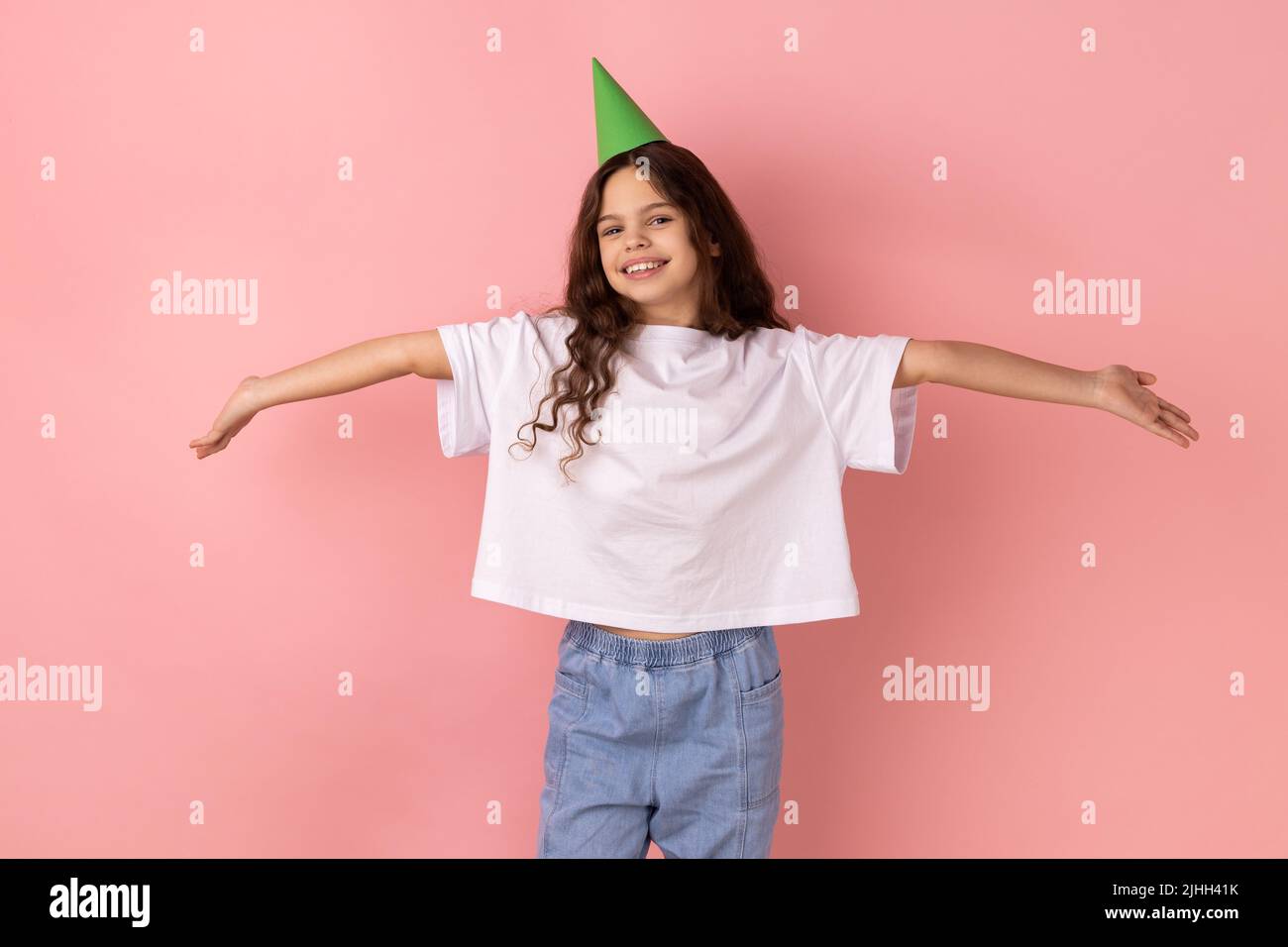 Portrait d'une petite fille satisfaite portant un T-shirt blanc en gardant les mains ouvertes, invitant, donnant un accueil chaleureux, saluant et partageant l'amour. Studio d'intérieur isolé sur fond rose. Banque D'Images