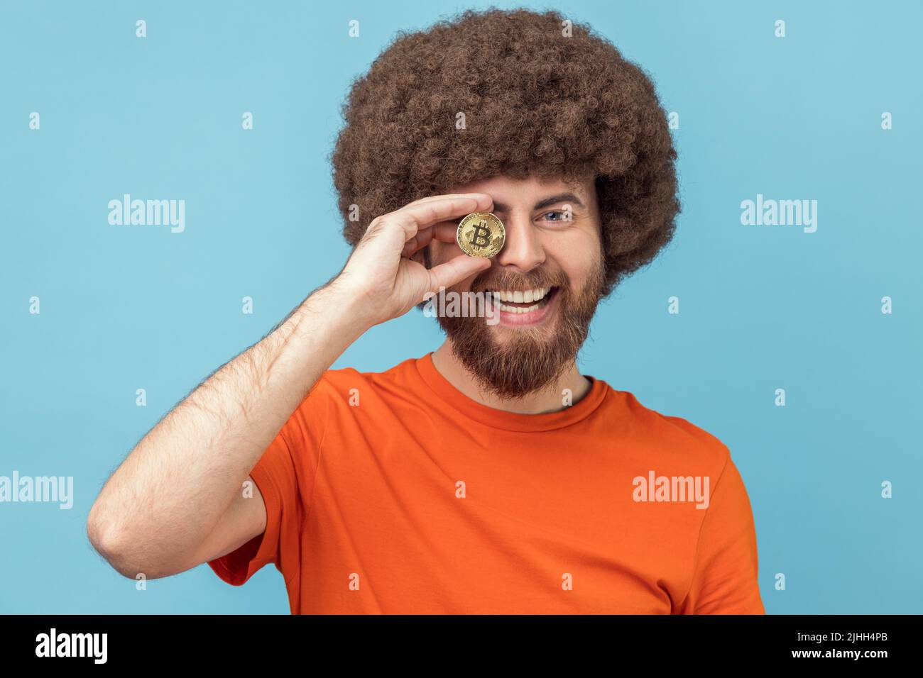 Portrait de l'homme optimiste avec la coiffure afro portant un T-shirt orange couvrant l'oeil avec le bitcoin d'or, ayant l'expression positive, la monnaie crypto. Studio d'intérieur isolé sur fond bleu. Banque D'Images