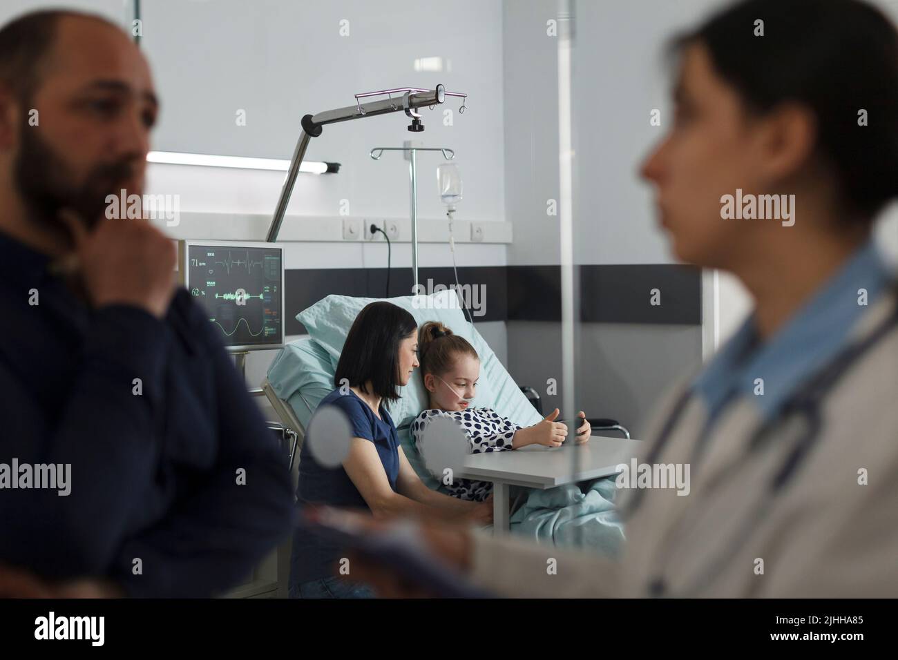 Fille malade jouant à des jeux sur smartphone avec la mère pendant que le père parle avec un pédiatre expert. Hospitalisé malade petit enfant regardant des dessins animés sur téléphone mobile tout en se reposant dans la salle pédiatrique de l'hôpital Banque D'Images