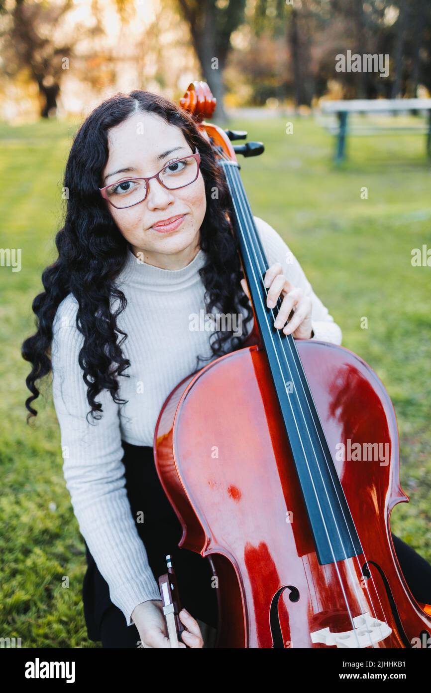 Bonne jeune femme brune avec des lunettes souriantes et jouant du violoncelle au coucher du soleil dans le parc. Verticale. Banque D'Images
