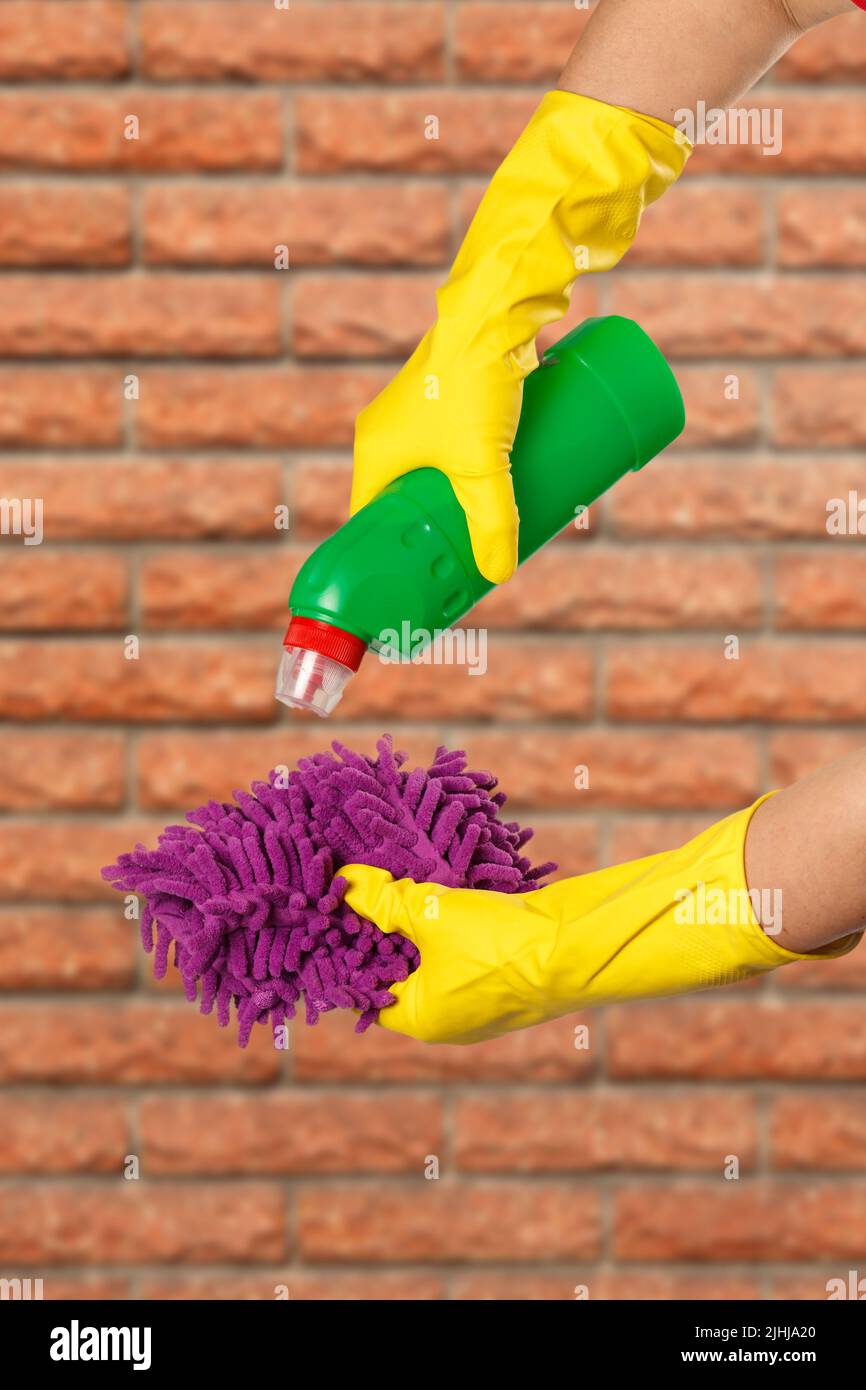 Les mains des femmes dans des gants de protection jaunes avec une bouteille de liquide vaisselle et un chiffon avec un mur de brique sur le fond. Concept de lavage et de nettoyage. Banque D'Images