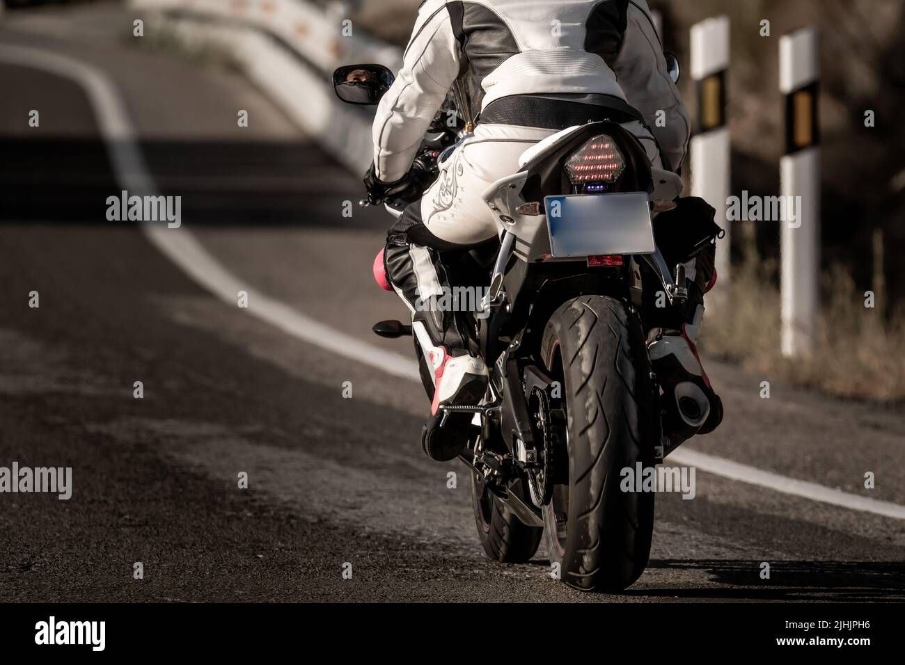 vue arrière d'une jeune motard vêtue d'un costume de protection en cuir et d'un casque de sécurité qui fait rouler sa moto sur une route asphaltée Banque D'Images